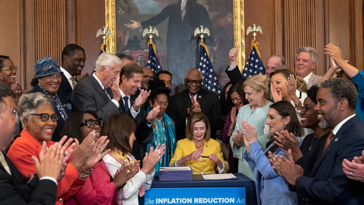 Nancy Pelosi sits at a table to sign the inflation reduction act. She's surrounded by people clapping.