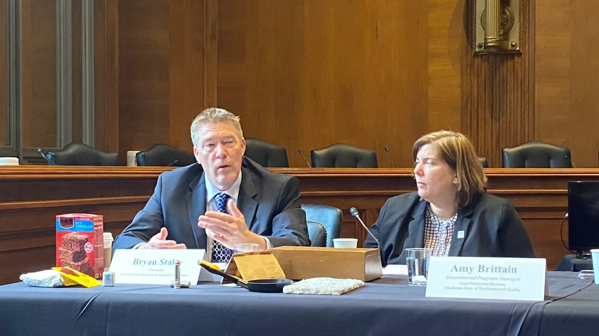 Two speakers sit at a table with product packaging that contains PFAS.