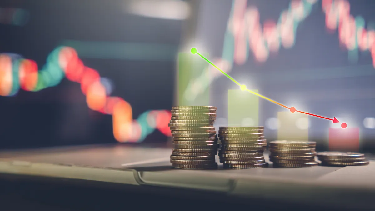 investment concept, stacked coins with a background monitor showing data graphs with an arrow pointing to a low point.