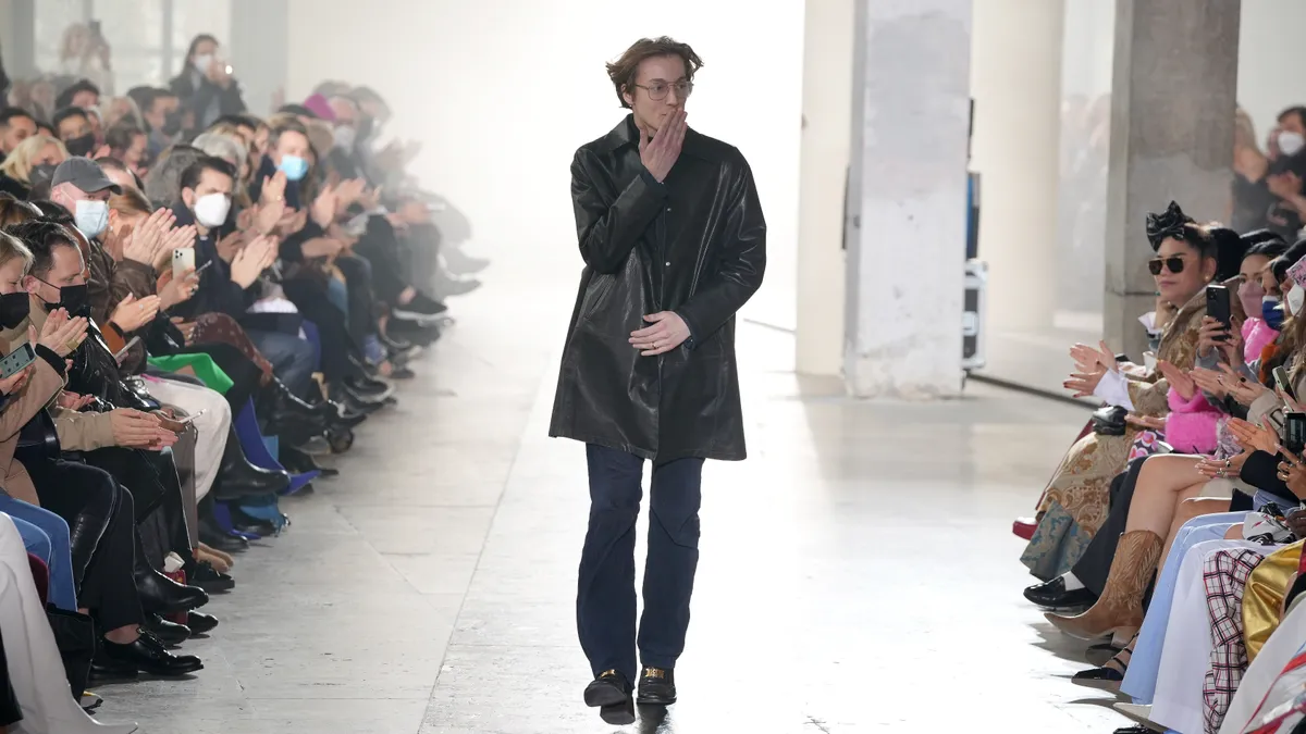 A man in a black coat blows a kiss to the crowd on a fashion runway