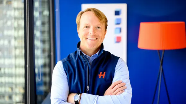 A person smiles for a portrait while wearing a blue vest with an "H" symbol on the chest in an office with blue walls.