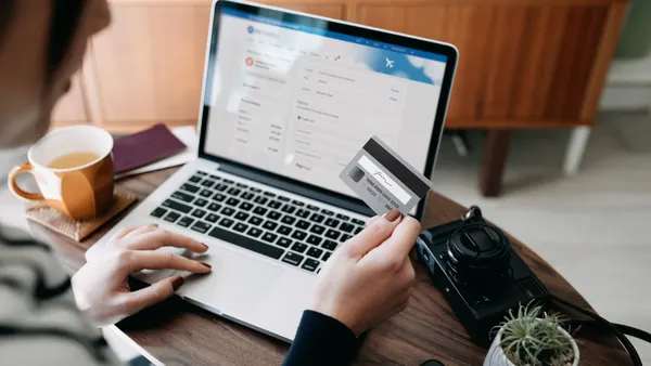 Over the shoulder view of young Asian woman shopping online for flight tickets on airline website with laptop, entering credit card details to make mobile payment at home. Camera and passport on the table. Travel planning. Booking a holiday online