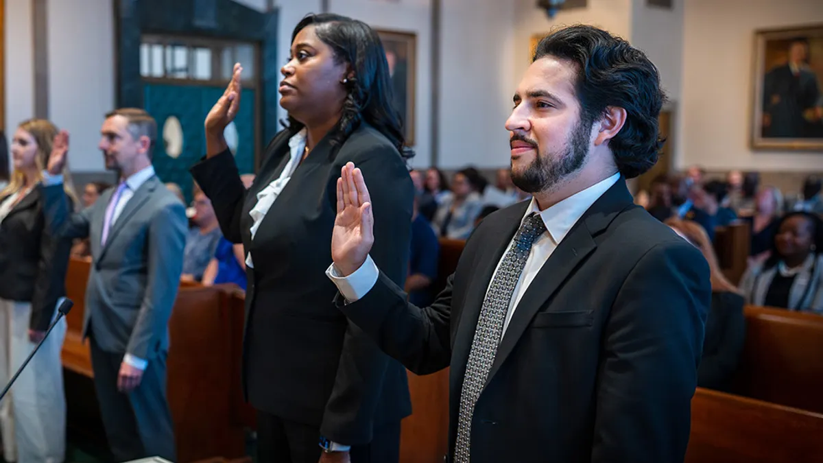 In a formal courtroom, three adults in business attire raise their right hands during an oath, with a diverse audience watching in the background.