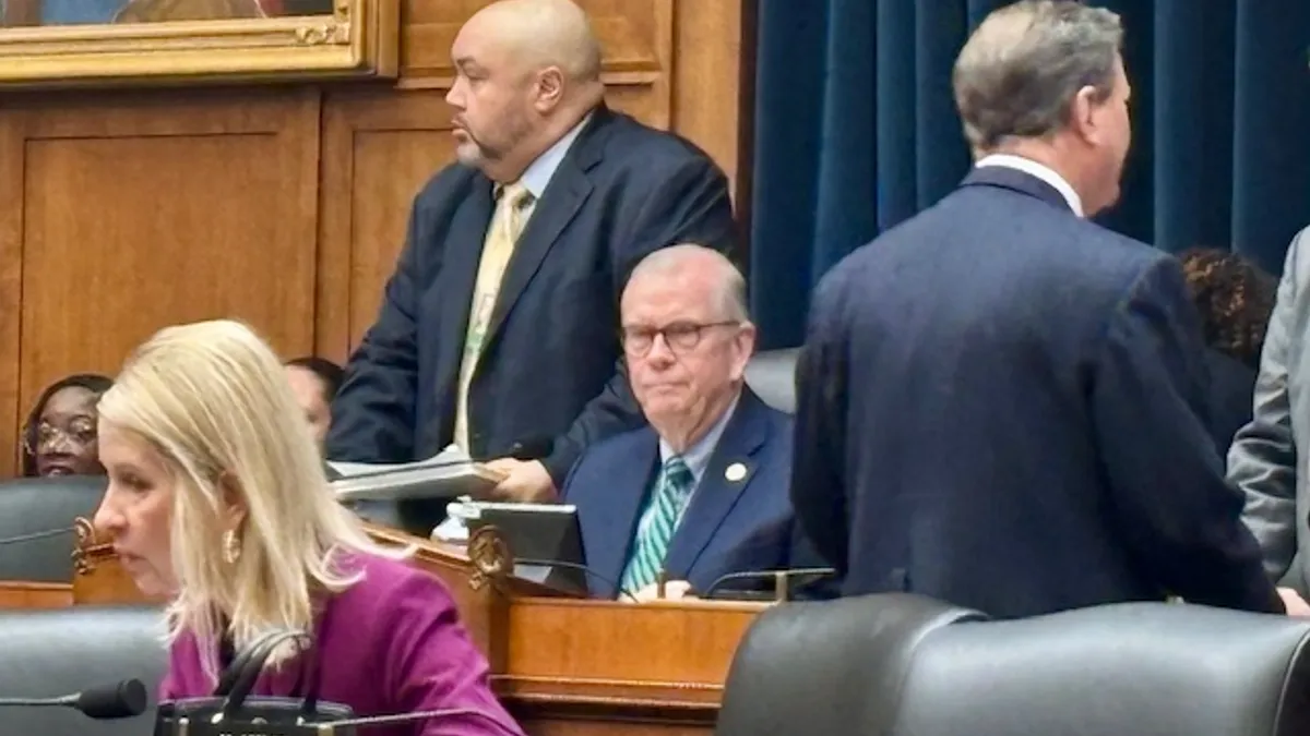 A man is sitting at a desk at a dias. Other people are sitting or standing around them.