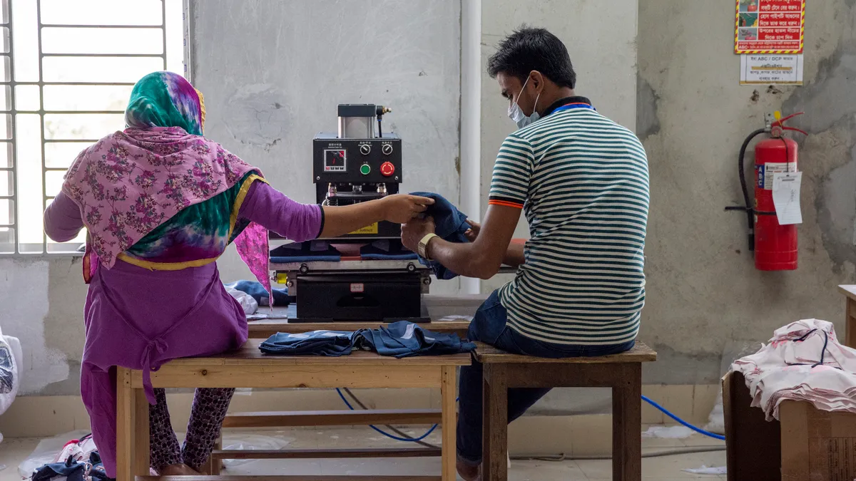 Two garment workers are seen from behind, working together on a piece of material.