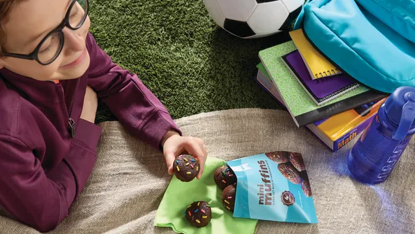 Kid with glasses wearing purple sweatshirt and sitting on grass with a bag of chocolate mini muffins with rainbow sprinkles.