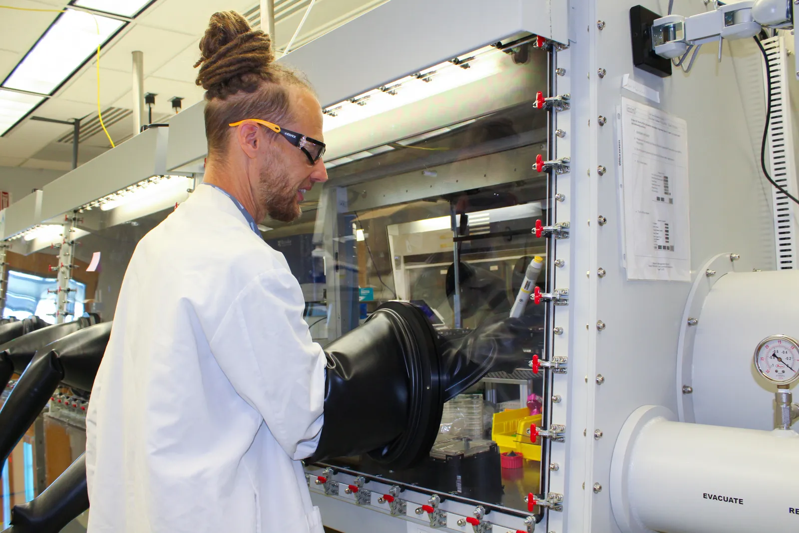 A person wearing a lab coat and goggles in a biotechnology lab performs experiments with their hands in a sealed chamber.