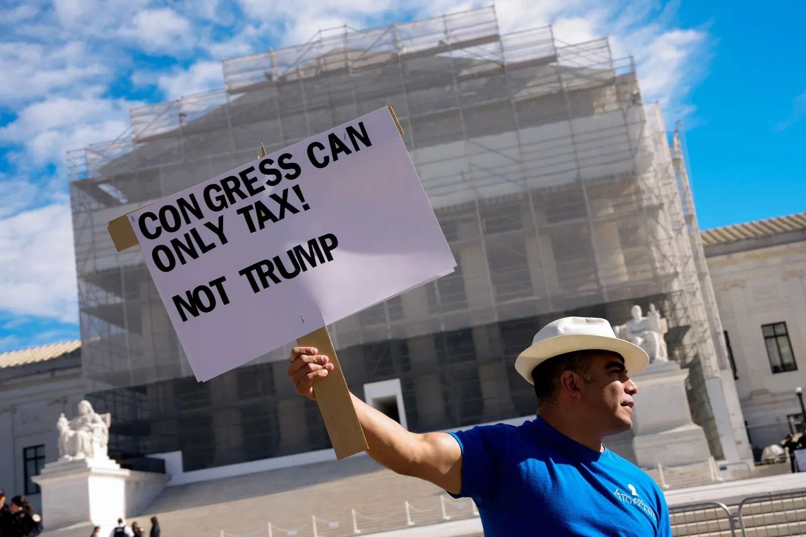 A person in a blue shirt stands in front of building under construction holding a sign that says “Congress can only tax! Not Trump.”