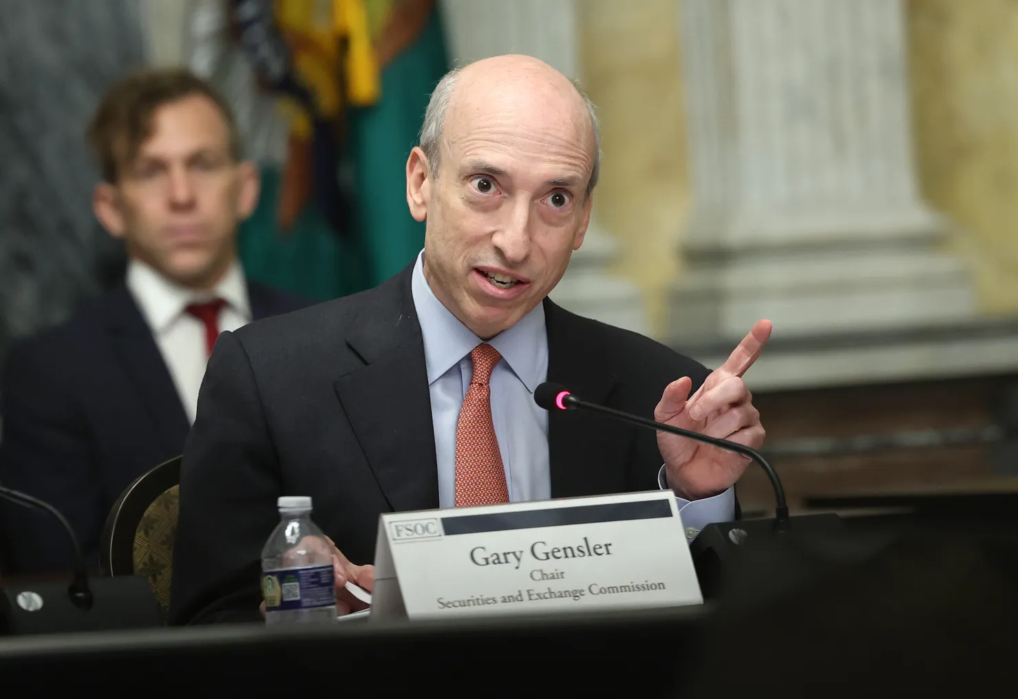 Gary Gensler is in focus speaking with a finger raised in front of a name card. An aide sits behind him out of focus.