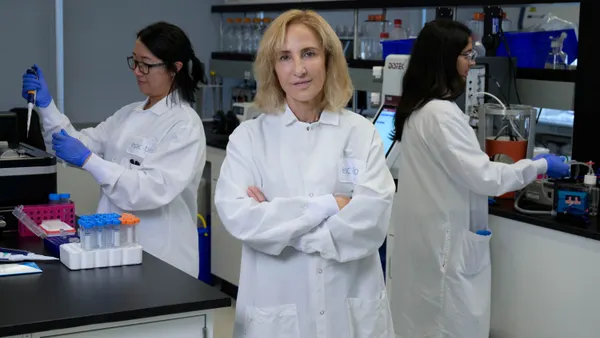 a photo of Epicrispr Biotechnologies CEO Amber Salzman standing in a white lab coat facing the camera with a woman behind her to her left in a white lab coat with black glasses holding a tube and syringe, and a woman in a white labcoat behind to the right facing the right touching lab tools.