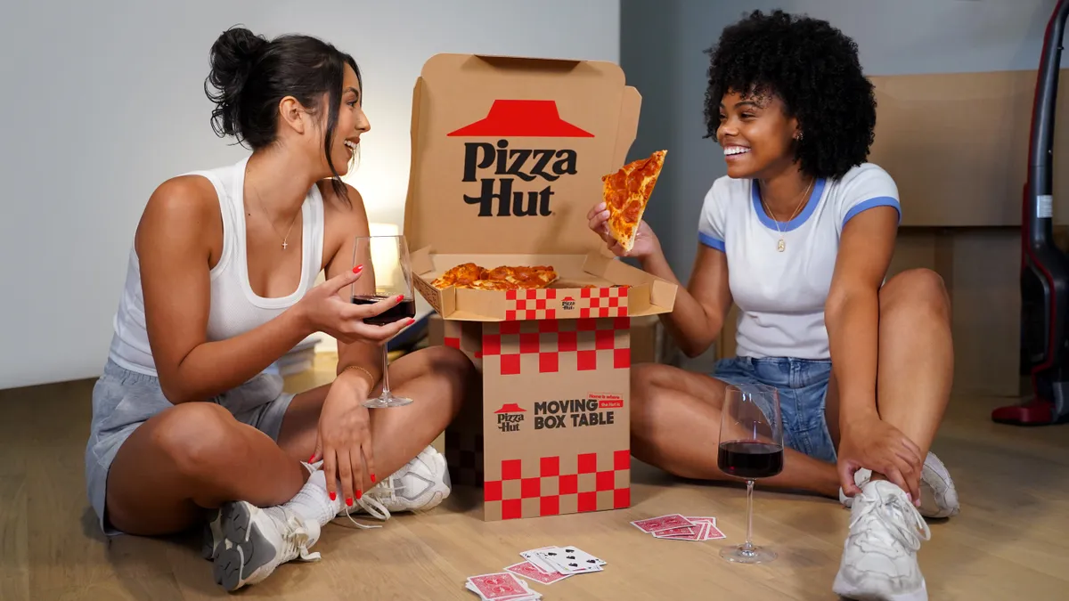 Two people eat pizza out of a Pizza Hut box which has a base that can be folded into a miniature table.