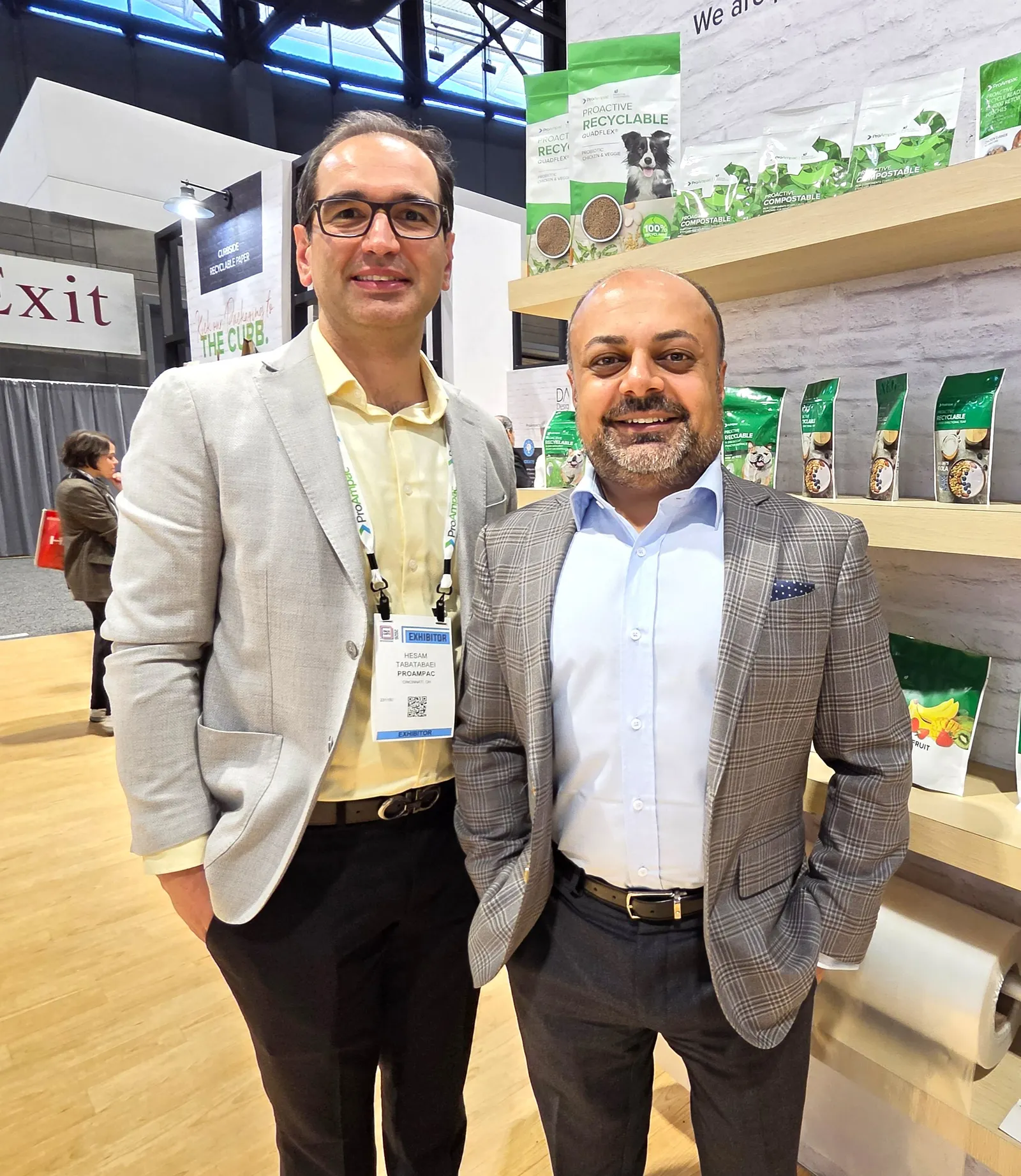 Two people stand in an exhibition hall booth with flexible pouches on shelves behind them.