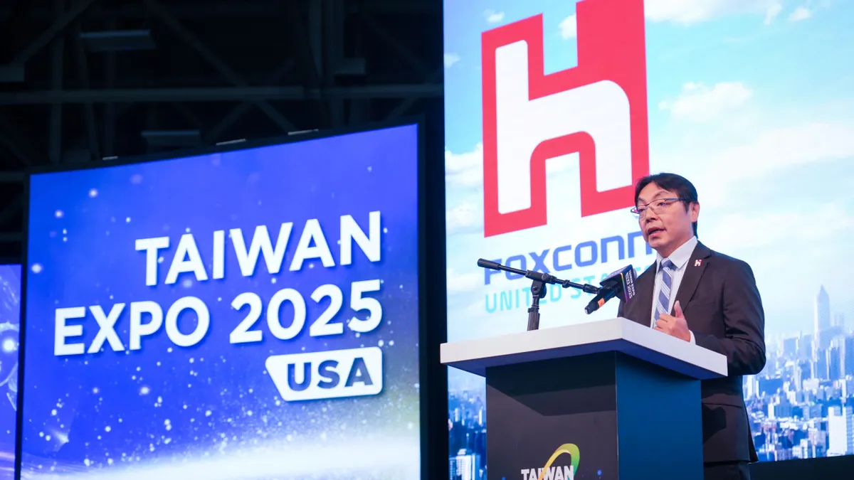 A dark-haired person with glasses speaking into a mic at a podium with the Taiwan Expo USA on the left of him.