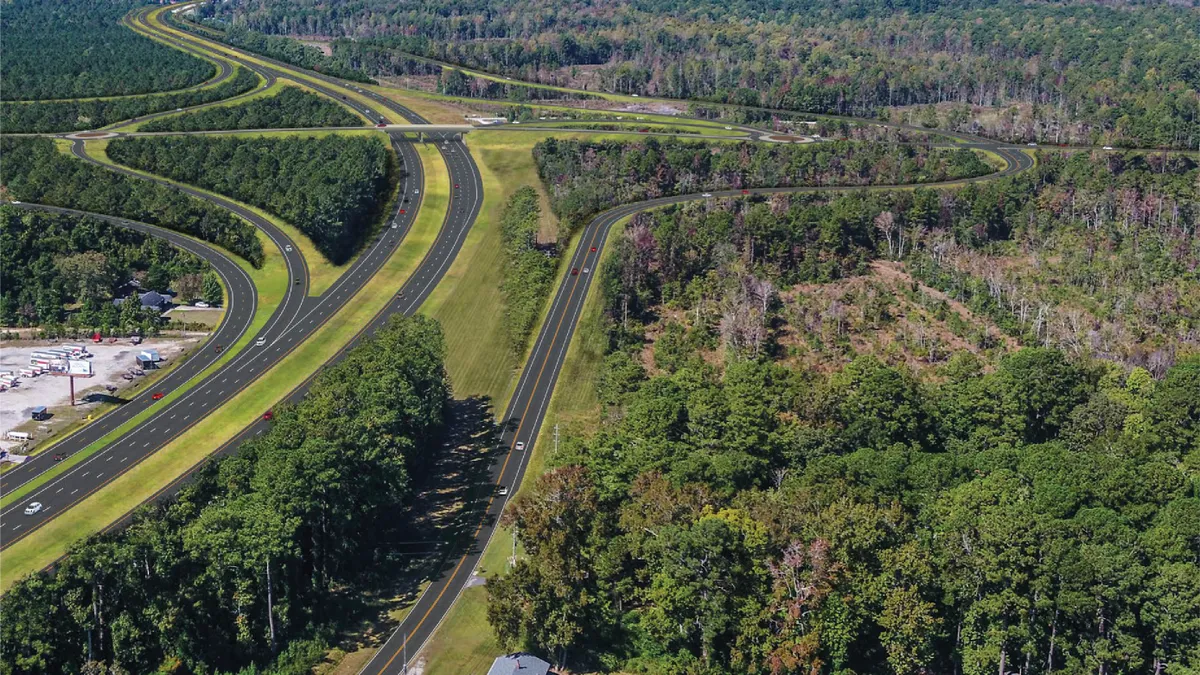 An aerial shot of a stretch of the U.S. 70 Highway over North Carolina. The stretch of highway winds on a clear day with blue sky, with a lot of green foliage near the roads.