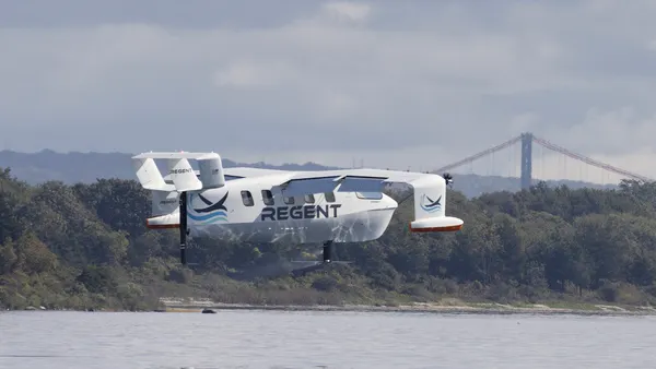 A winged white aircraft flies closely above a sea with tree-lined coast behind.
