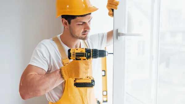 Handyman in yellow uniform installs new window by using automatic screwdriver.