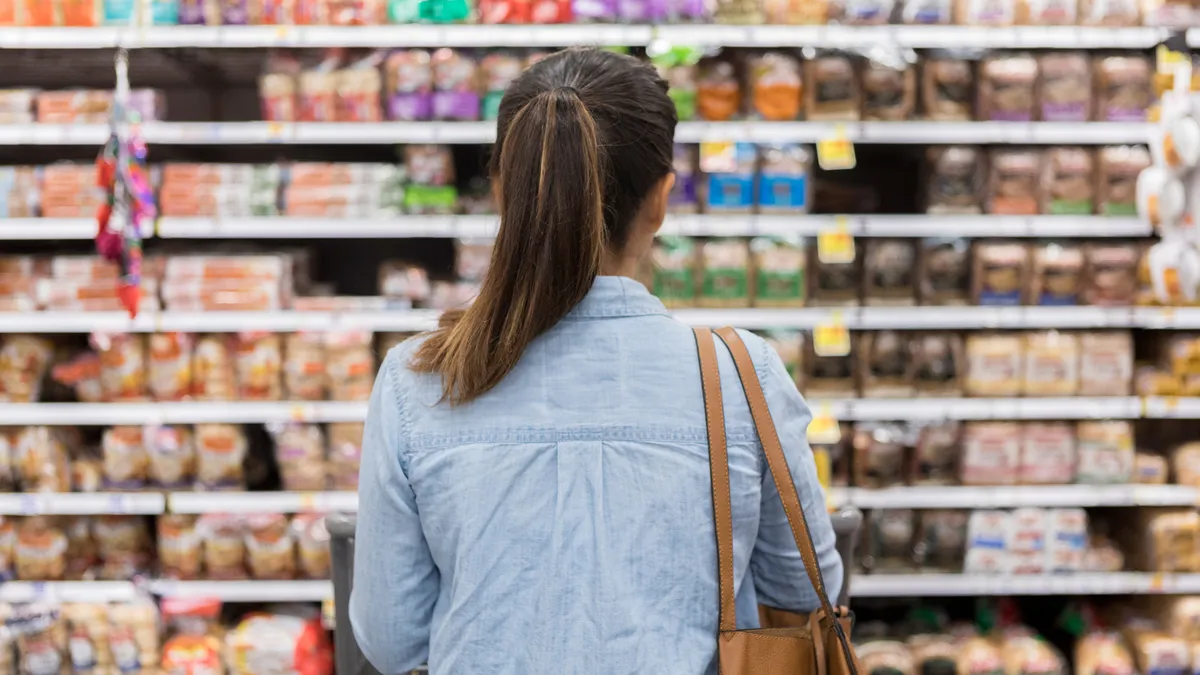 In this rear view, an unrecognizable woman stands with a shopping cart in front of a shelf full of food in the bread aisle of a grocery store.