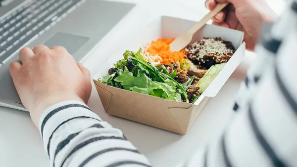 A photograph of a person's arms holding a wooden knief that is resting in a small paper box of food.  Another hand is on a gray latop.