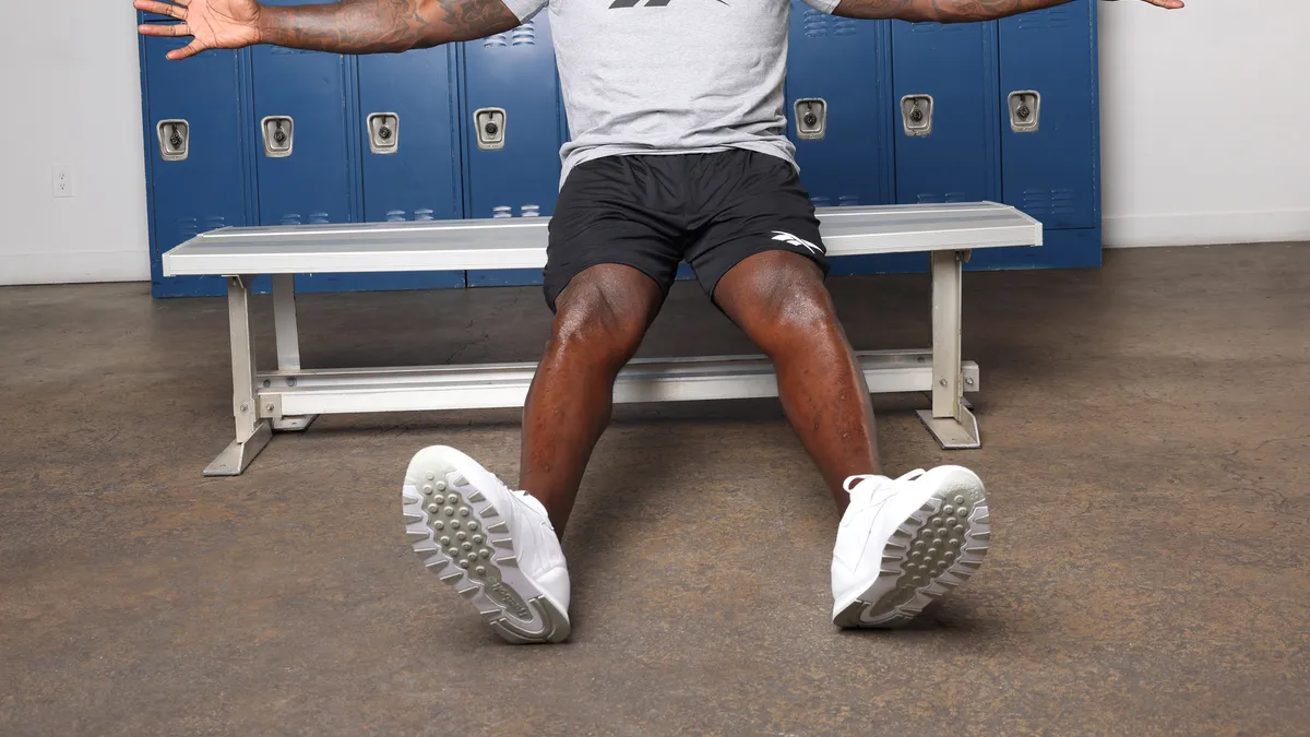 Shaquille O'Neal sitting on a bench in a Reebok shirt with his arms flung wide.