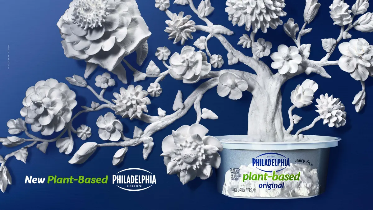 A white tree with long branches and large flowers appears to be growing out of a tub of Philadelphia Plant-Based spread, which is on a blue background.