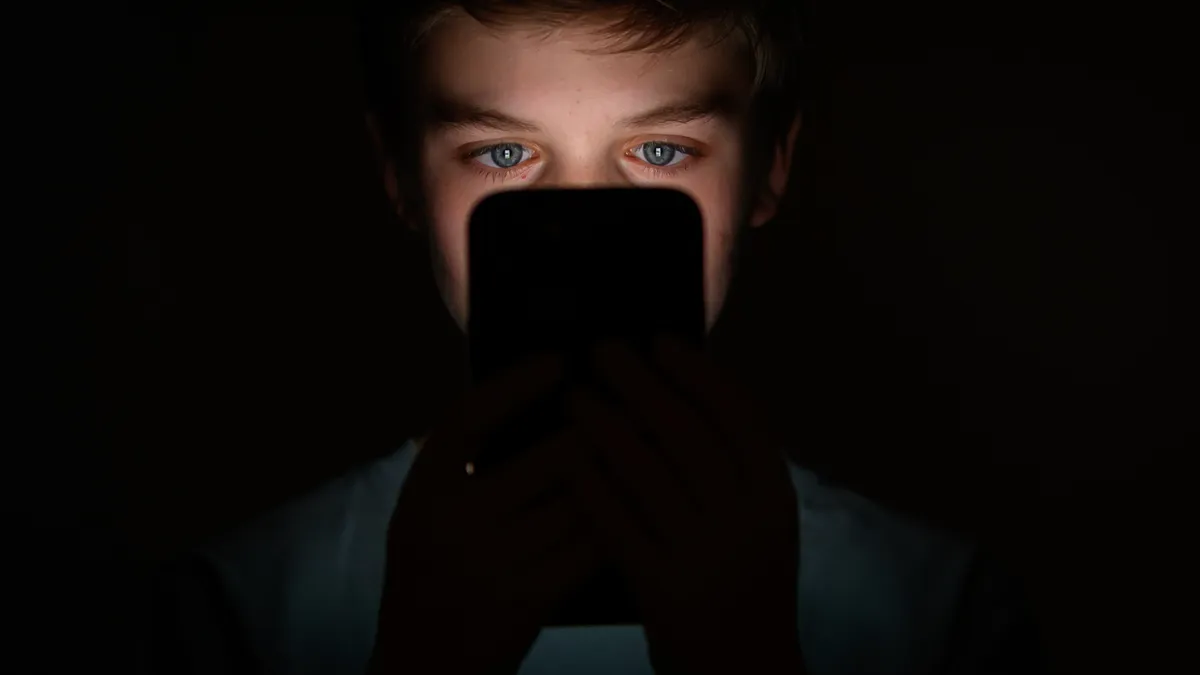 A teenager looks into a cellphone screen in the dark as the phone's light illuminates their face.