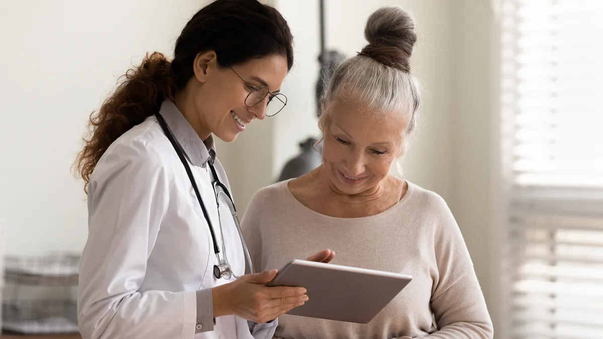 Happy female doctor and senior patient look at tablet screen discuss treatment.