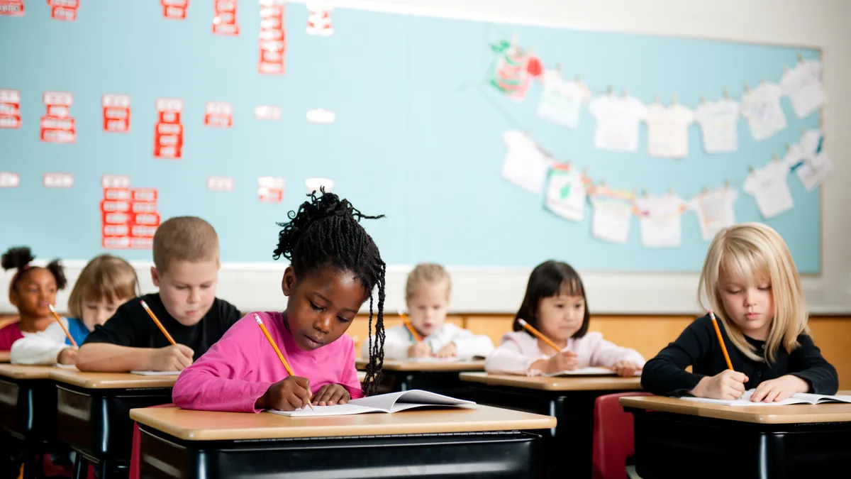 A handful of students are sitting at individual desks in a classroom. ALl the students have pencils in their hands and are writing in notebooks.