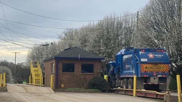 A garbage truck with Republic Services branding sits on a platform next to a small brick building on a dirt road.