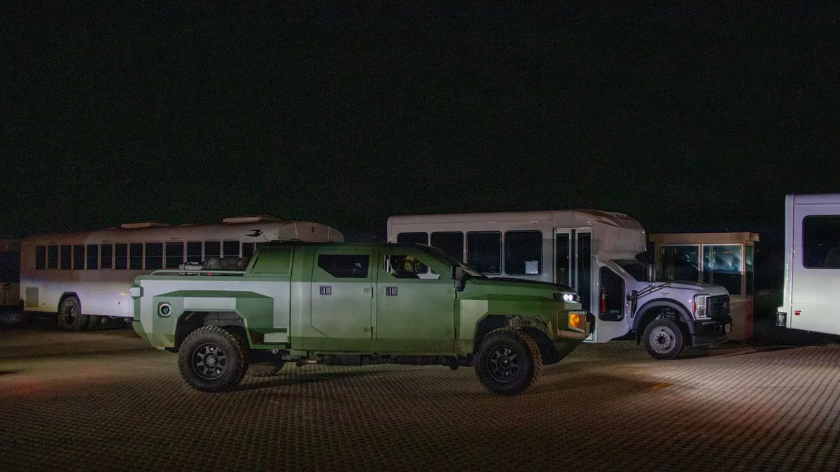 A jungle green military truck in the middle at night, with three white buses in the background.