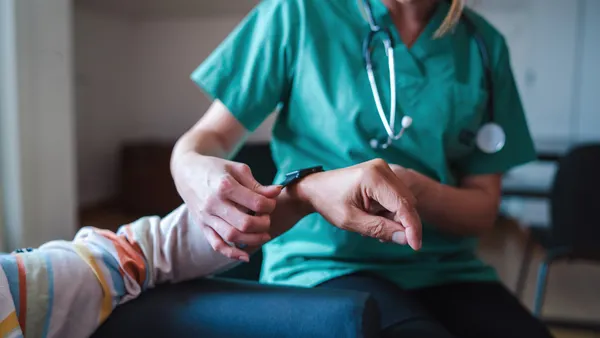 A nurse in green scrubs puts a smart watch on a patient in a grey, blue and orange striped shirt.