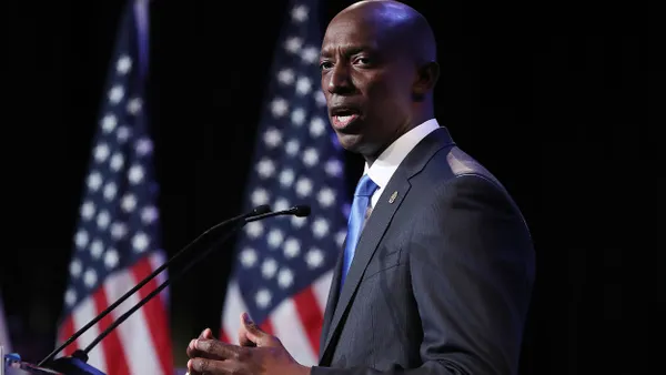 A person wearing a suit standing at a podium with two U.S. flags behind.