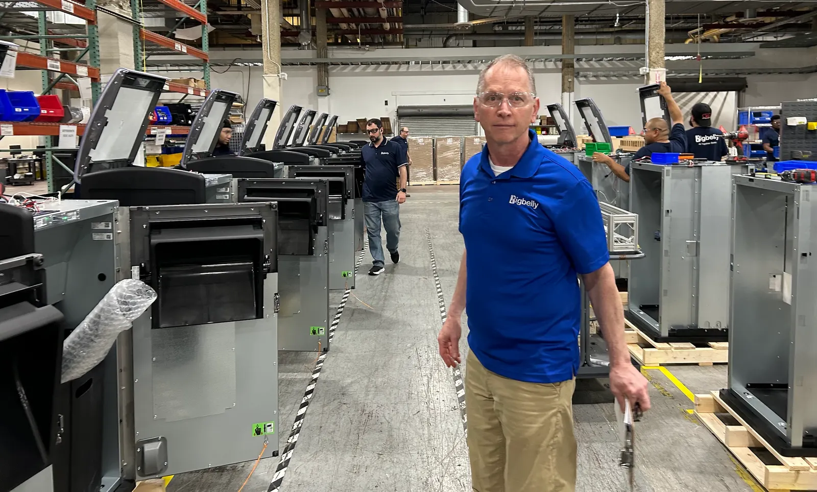 A person in a Bigbelly shirt and safety glasses stands in an assembly line for trash bins