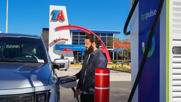 A photo of a person plugging an EV charging plug into a vehicle. There is a building in the background with signs that say "TA," "Welcome" and "Jacksonville South travel center." The EV charger has wording that reads "BP Pulse."