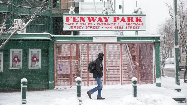 A person wearing a heavy jacket carrying a backpack walks through snow in front of a sign for Fenway Park.