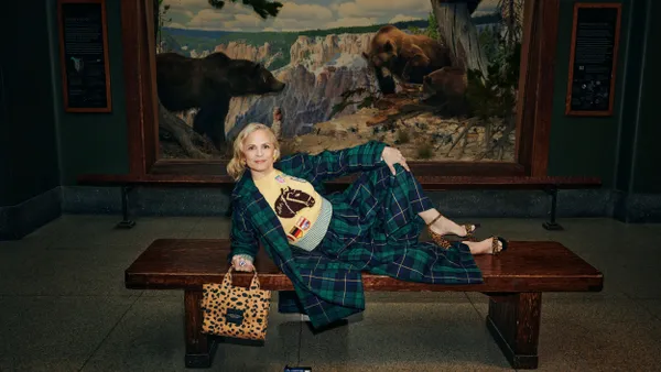 A person reclines on a bench in front of a large painting of three bears. They are wearing a plaid green outfit with a bright yellow sweater and leopard print heels that match their bag.