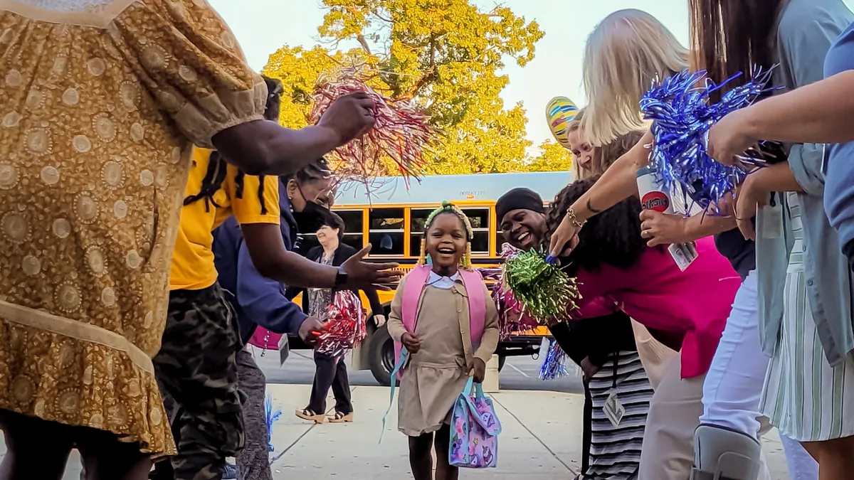 A young student walks between two lines of people holding pom-poms and high-fiving. A yellow school bus is in the background