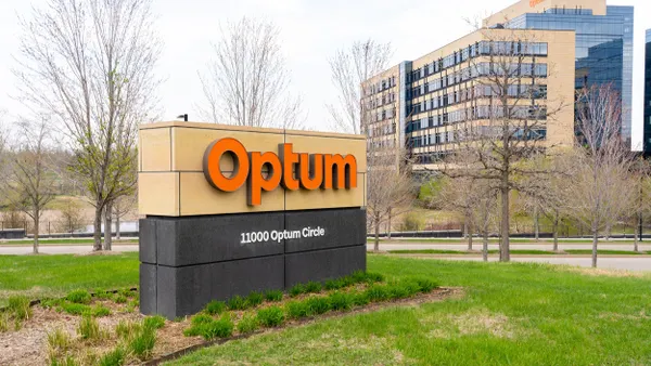 A tan and black monument sign with bold orange "Optum" lettering and the address "11000 Optum Circle" sits on a grassy lawn with low shrubs in front of a large, modern office building with glass windows and beige panels. Bare trees and a cloudy sky are in the background.
