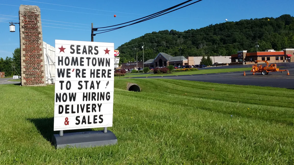 A bright white sign with two red stars, posted on green grass, promises "Sears Hometown we're here to stay."