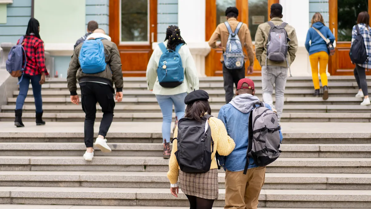 On a cold day the students are climbing up the steps towards the university entrance, everyone wearing coats and carrying backpacks containing their education books.