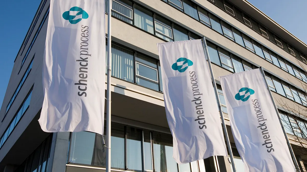 Four white rectangle-shaped Schench Process flags hung vertically in front of a five-story building.