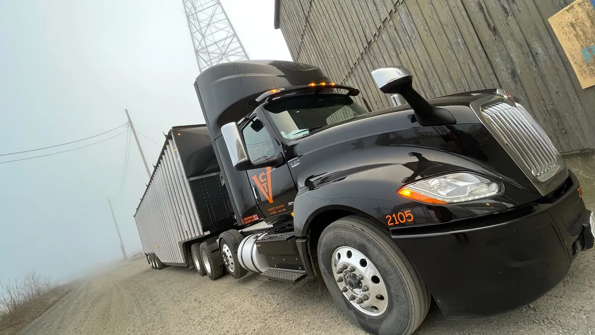 A black Veneer Chip Transport truck is parked on a gravel road next to a wooden fence.