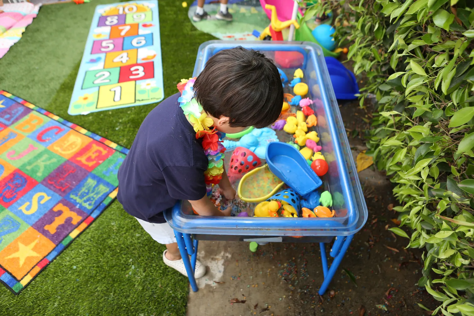 A small child reaches into a blue plastic bin of brightly colored toys on a sunny day during a kindergarten readiness summer learning program.