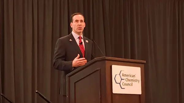 A man in a black suit and red tie speaking into a microphone, standing behind a podium.