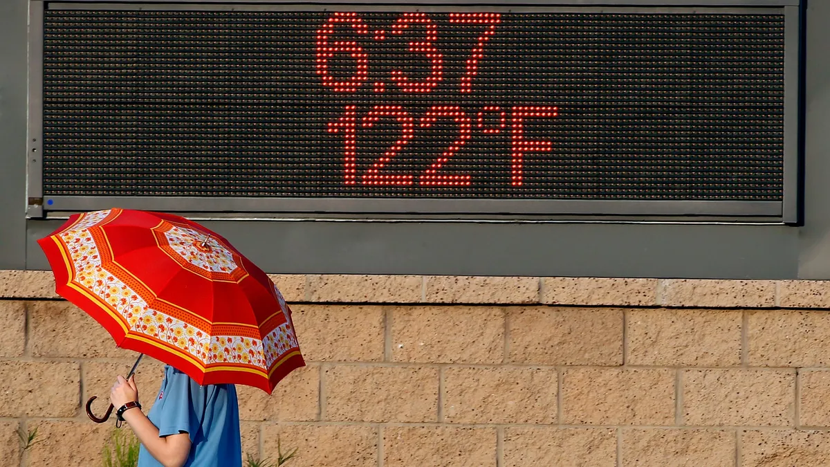 A person holds an umbrella in front of a digital sign reading 6:37 and 122 degrees Fahrenheit.