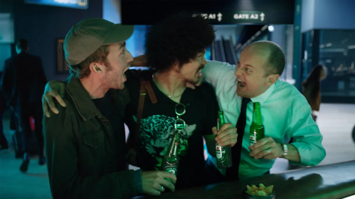 Three men with with their arms over each others shoulders while holding Heineken's.