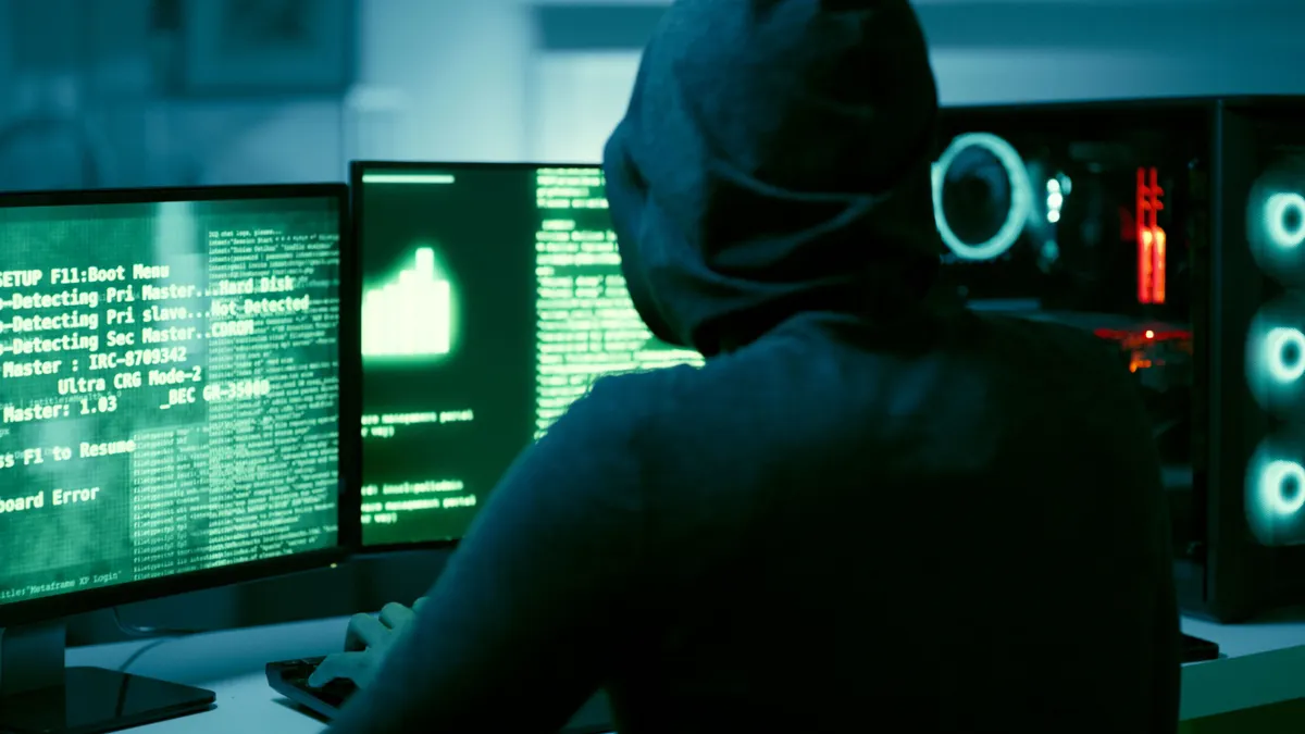 Person in a black hooded sweatshirt sitting in front of computer monitors displaying code.