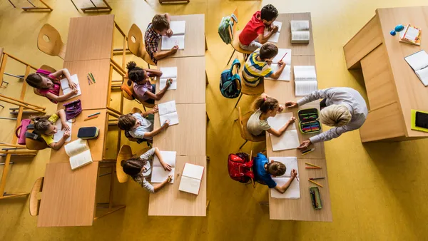 A view from above of an elementary classroom with rows of desks and students sitting at the desks with notebooks on the desks. An adult is standing in front of the desks helping a student.