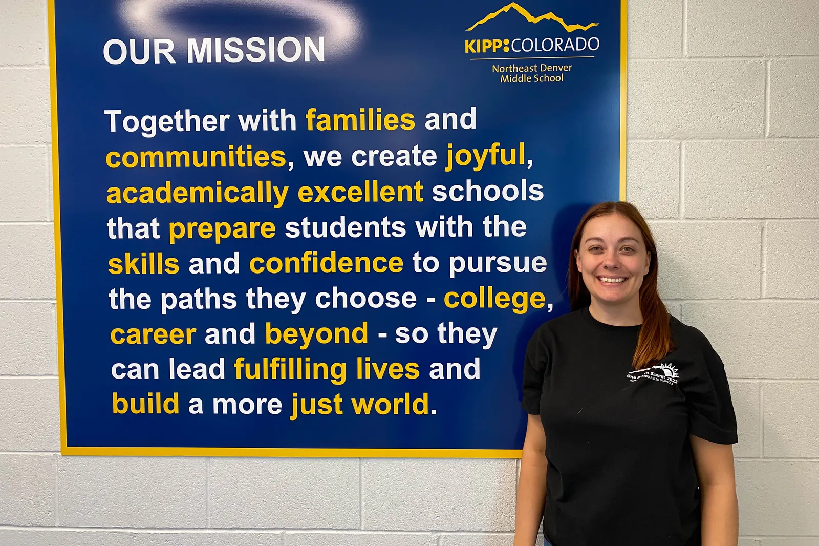 A person smiles in front a sign that reads "Together with families and communities, we create joyful, academically excellent schools that prepare students with the skills and confidence to pursue..."