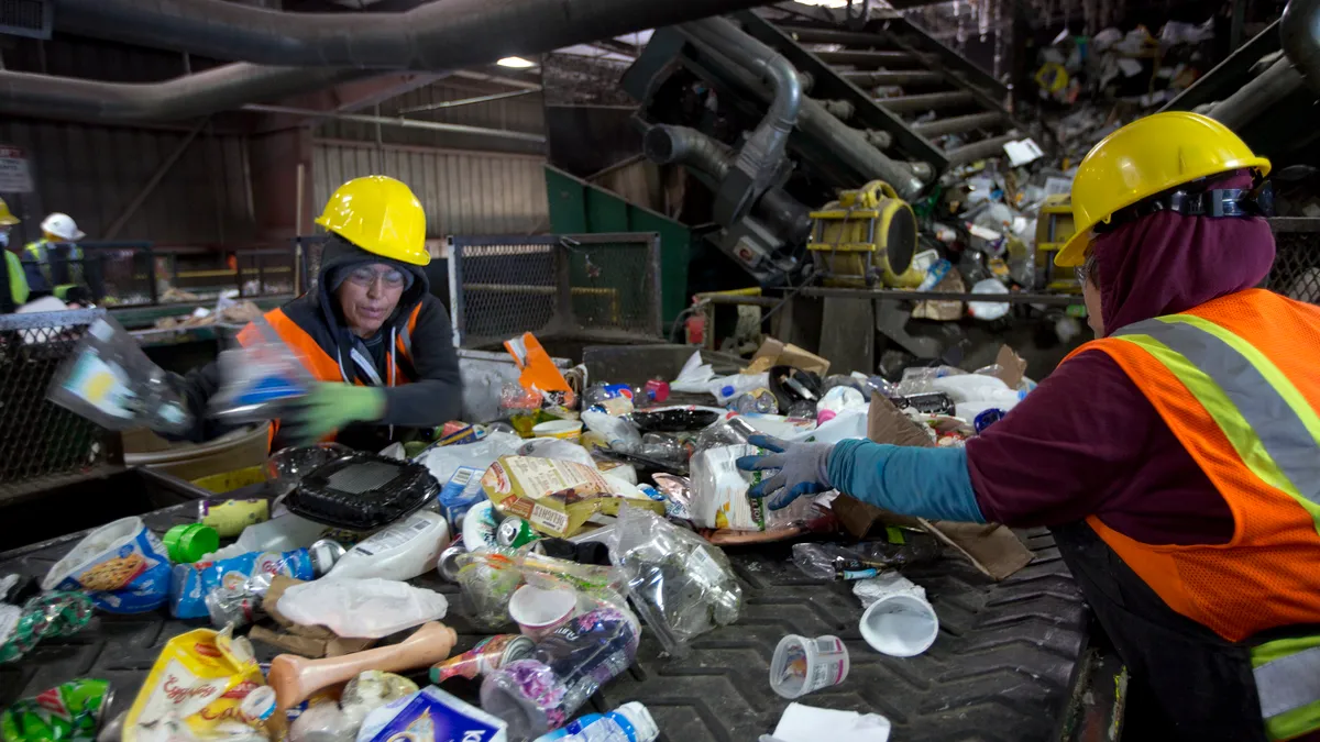 Two workers in yellow hard hats stand over a conveyor belt full of recyclable materials, primarily plastics, in an industrial facility