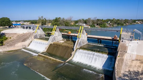 On a bright day, water flows over a dam and down into a larger body of water.